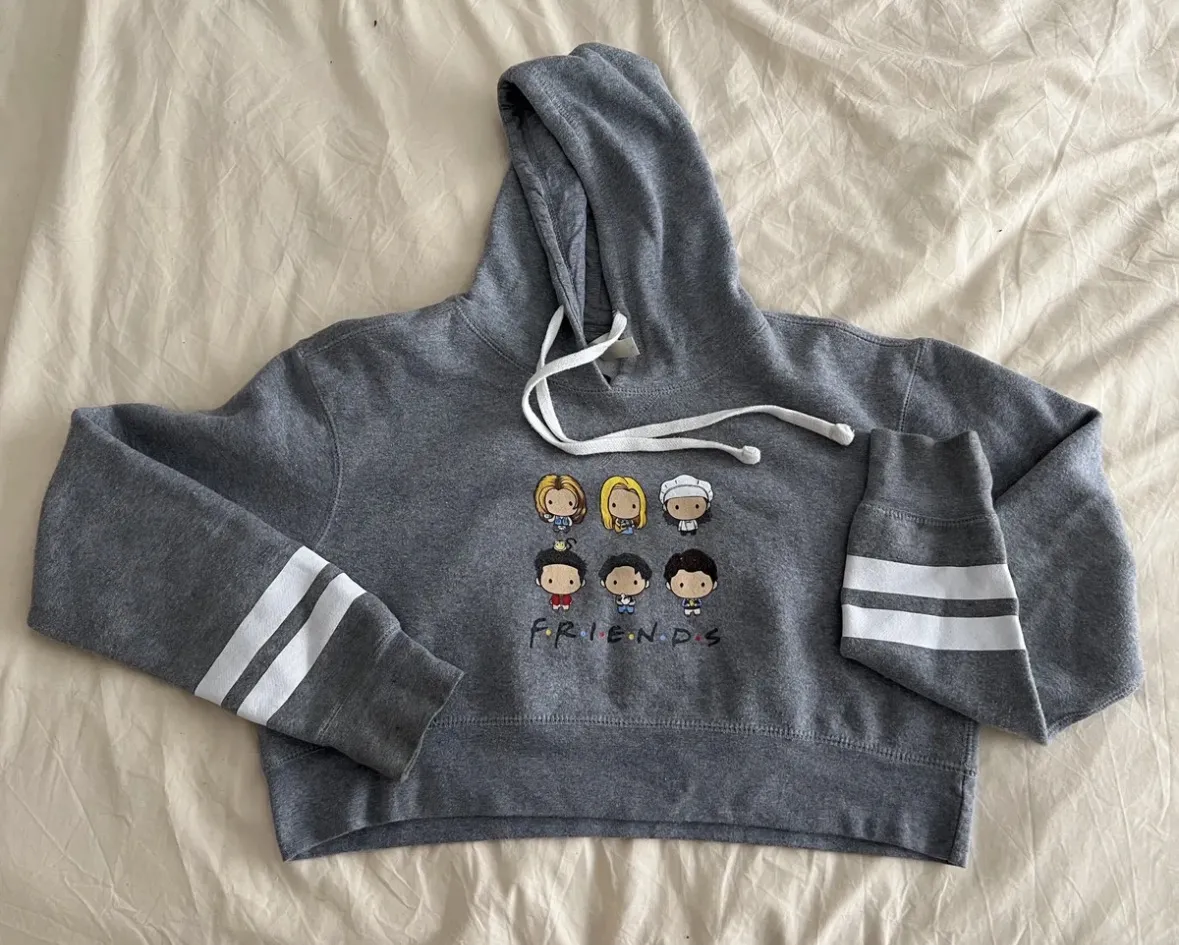 Importado. Original. Buzo crop gris con capucha, estampado de los personajes de la serie "Friends" y el logo de la serie. Tiene detalles de franjas blancas en las mangas y puños. Ideal para fans de la serie y para un look casual y moderno.