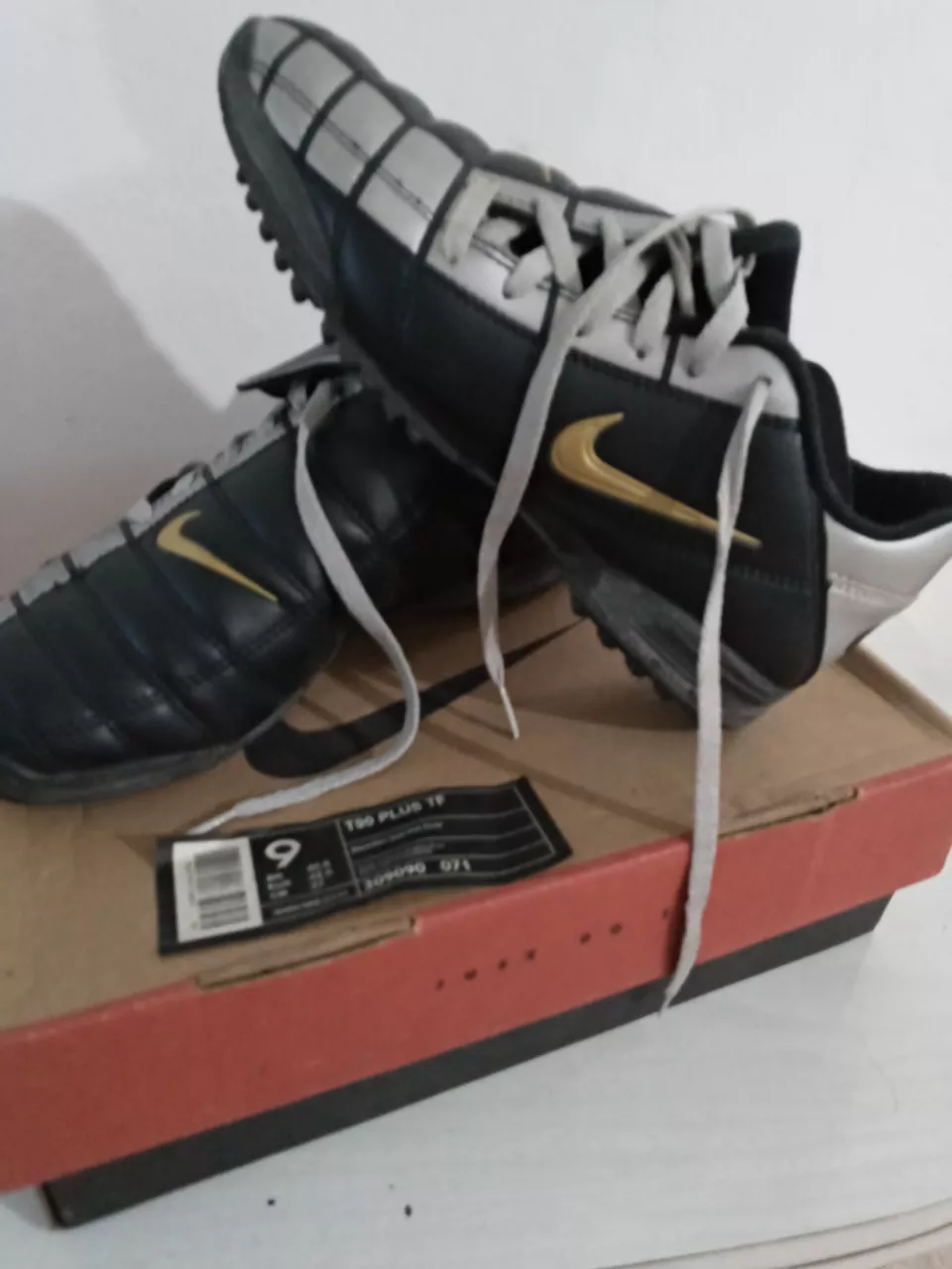 Botines de fútbol Nike T90 Plus TF, color negro y plateado con detalles dorados. Cordones grises. Suela con tapones para césped artificial.