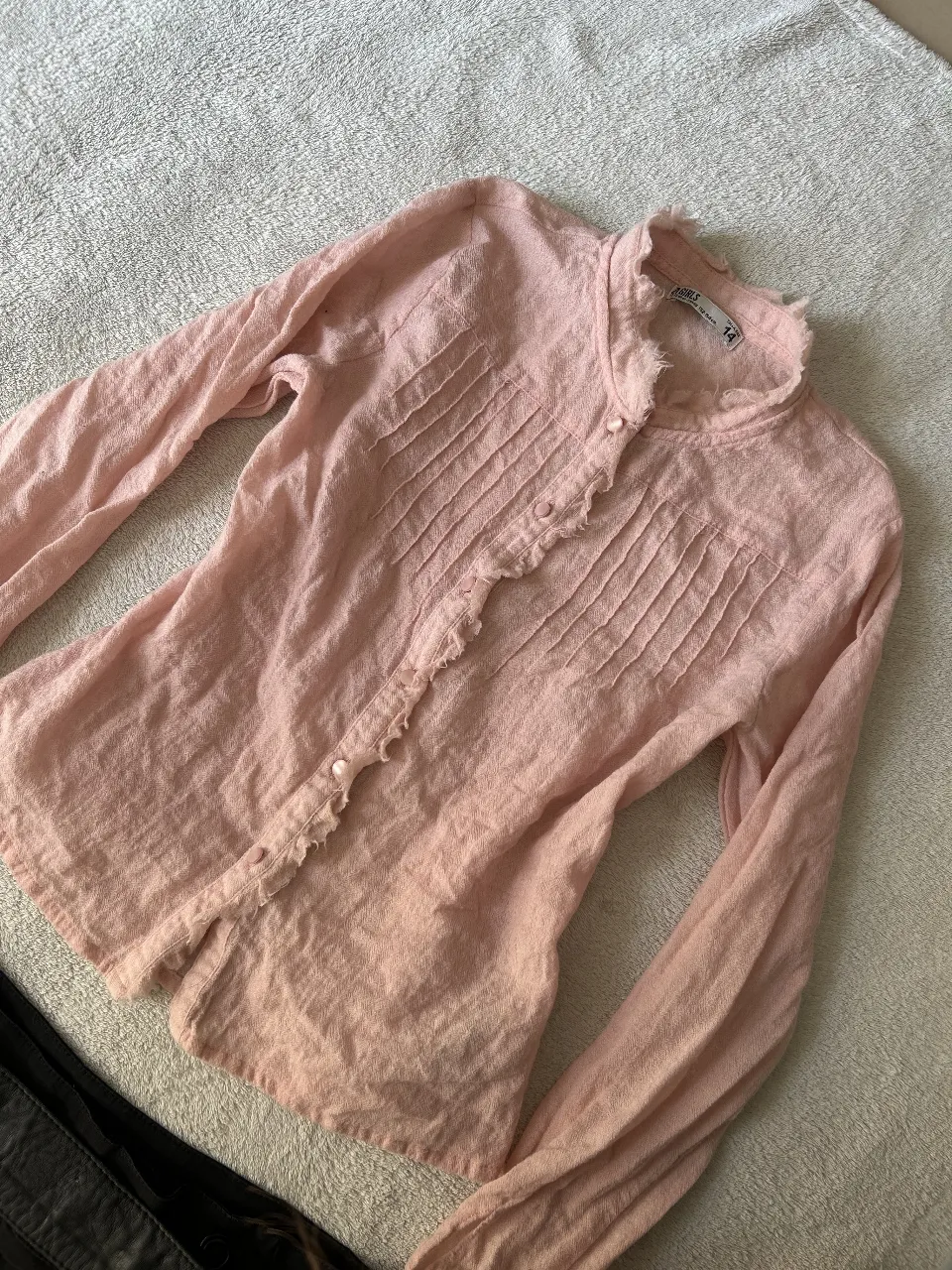 Camisa rosa de manga larga con detalles de frunces y volados en el cuello y la botonera. Posee botones nacarados y un diseño delicado, ideal para un look casual y femenino.