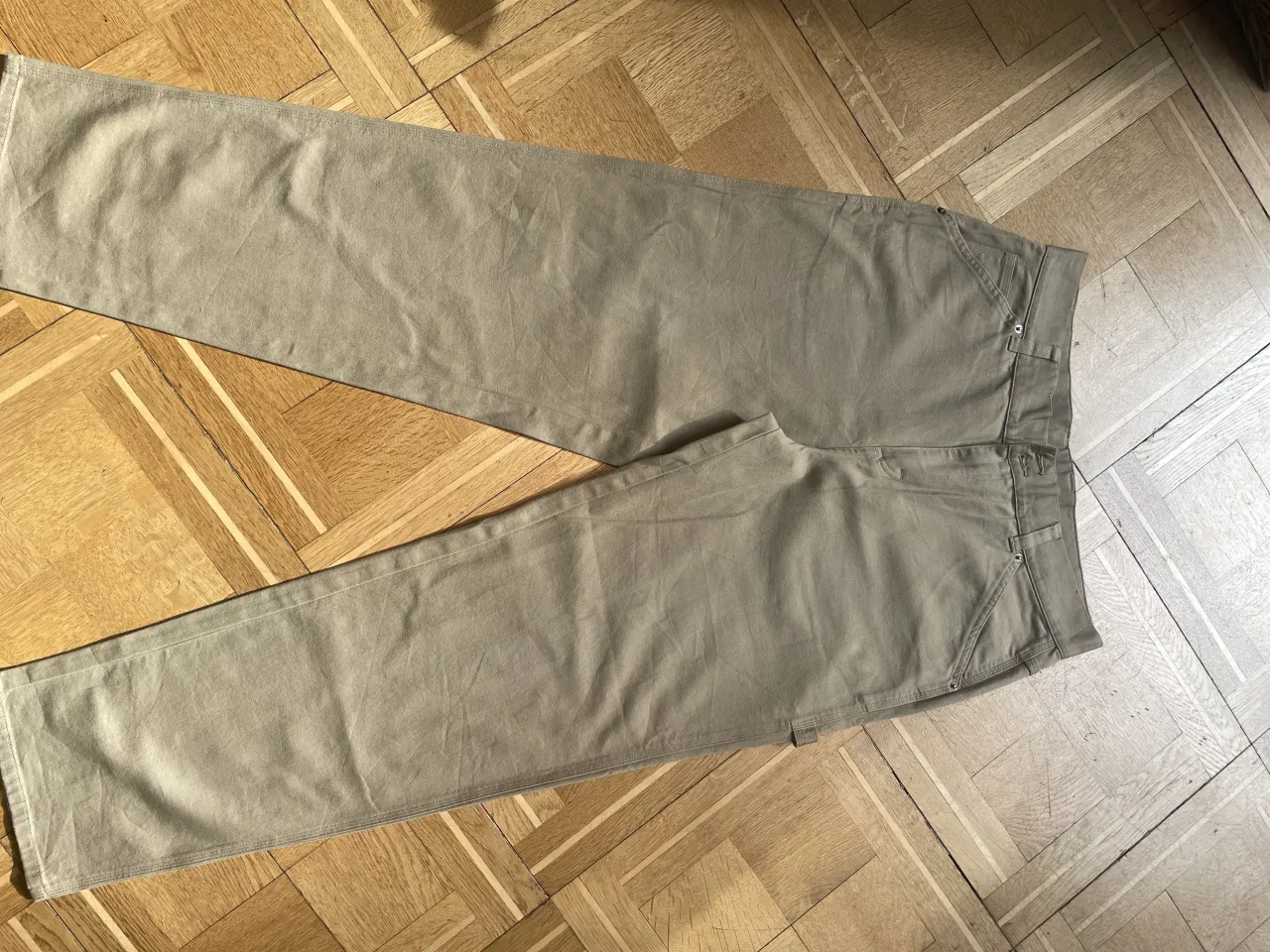 Pantalones de gabardina color beige. Tienen bolsillos frontales y traseros. Detalle de bolsillo adicional en el lateral derecho.