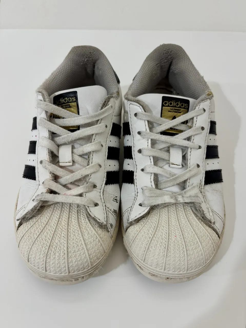 Zapatillas blancas y negras de Adidas Superstar. Tienen cordones blancos y las tres tiras negras características de la marca a los costados. La suela es blanca y la parte inferior es de goma.