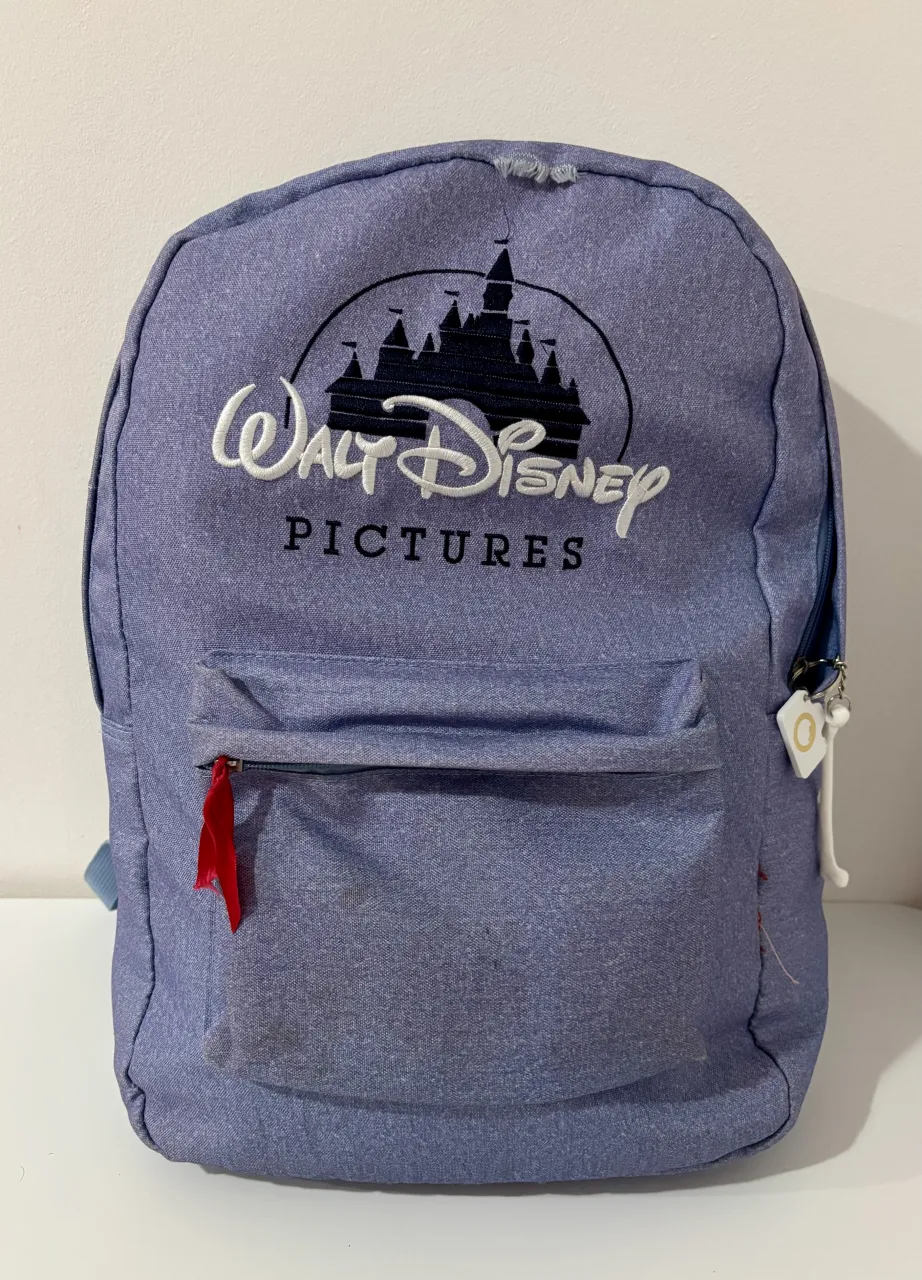 Mochila de tela color lila con logo de Walt Disney Pictures bordado en la parte frontal. Posee un bolsillo exterior con cierre. Las correas son acolchadas y ajustables.