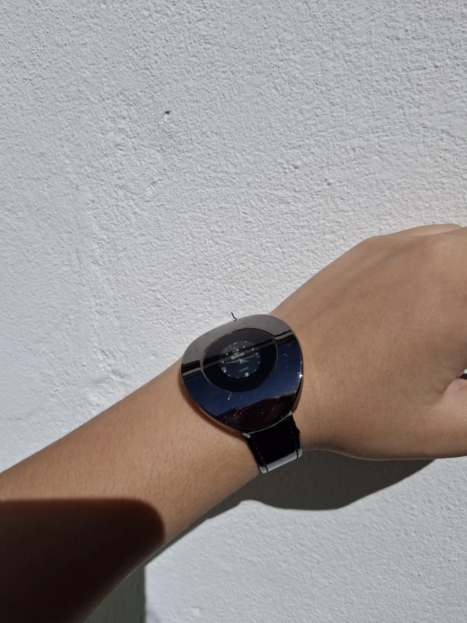 Reloj de pulsera con malla negra y caja metálica plateada de forma irregular. La esfera es negra con detalles plateados.