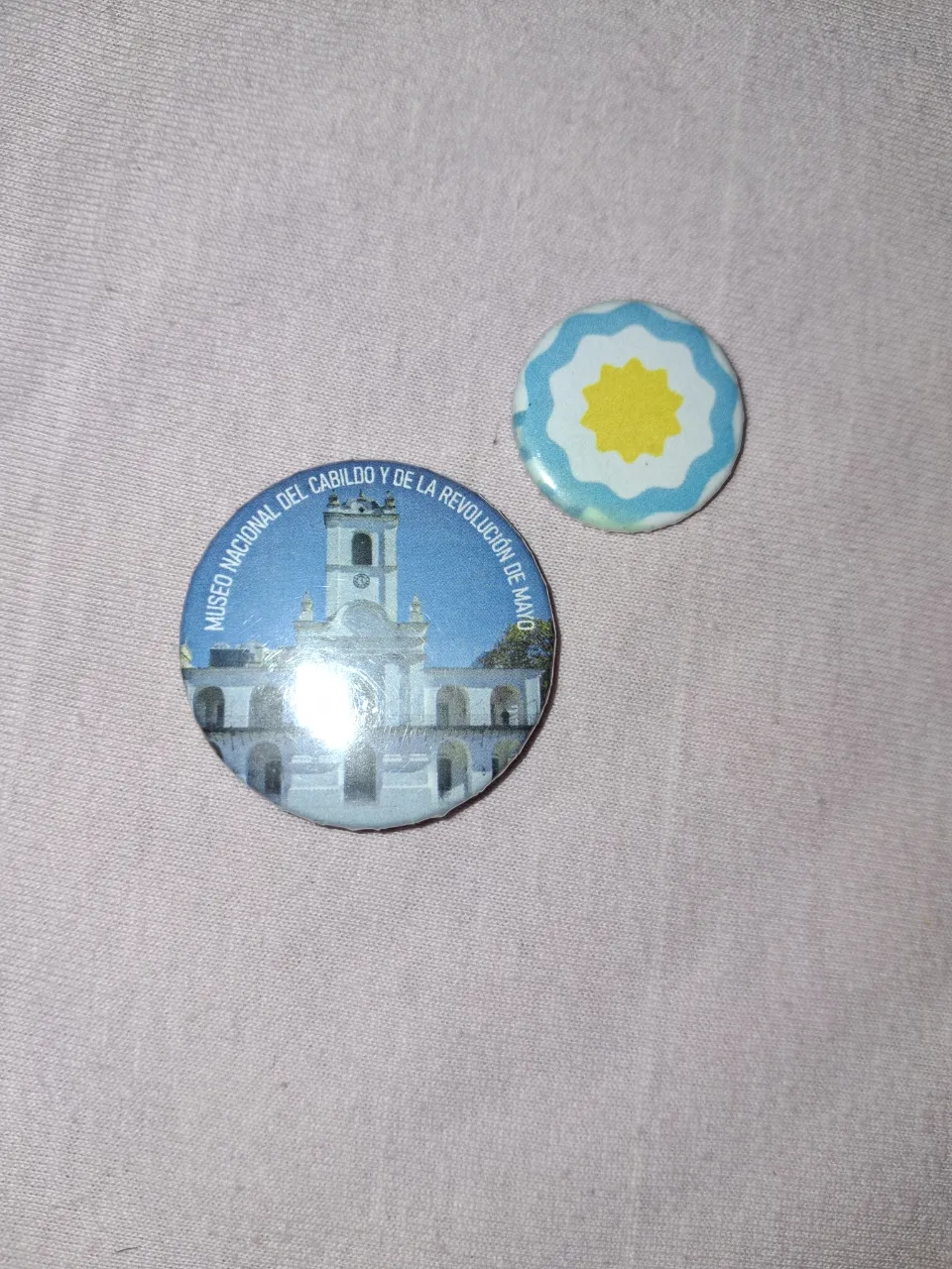 Lote de dos pines redondos. Uno muestra el Museo Nacional del Cabildo y de la Revolución de Mayo. El otro pin tiene el diseño de la escarapela argentina.