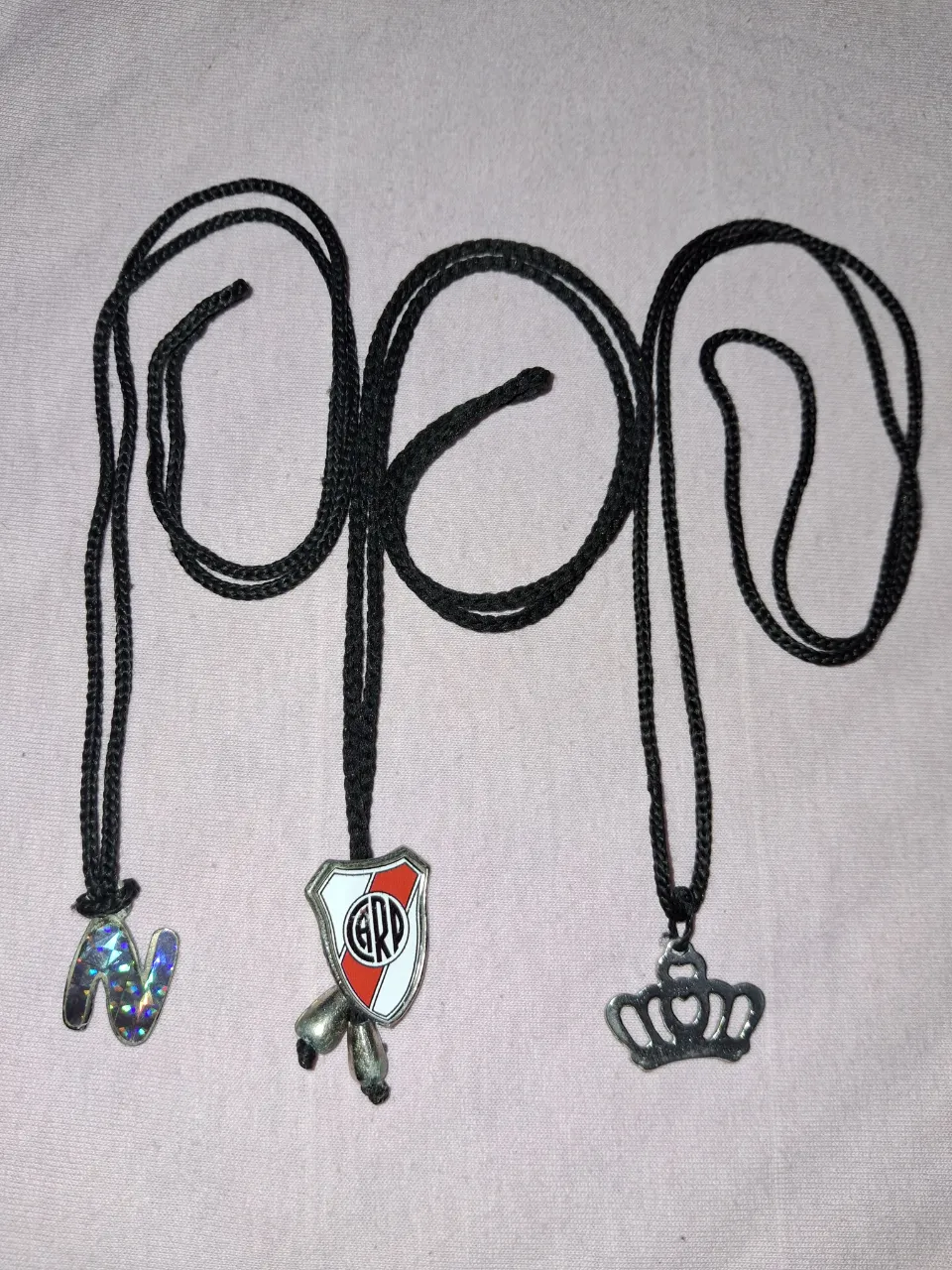 Lote de tres collares con dijes. Uno con la letra 'N' holográfica, otro con el escudo de River Plate y dos bolitas plateadas, y el último con un dije de corona plateada.