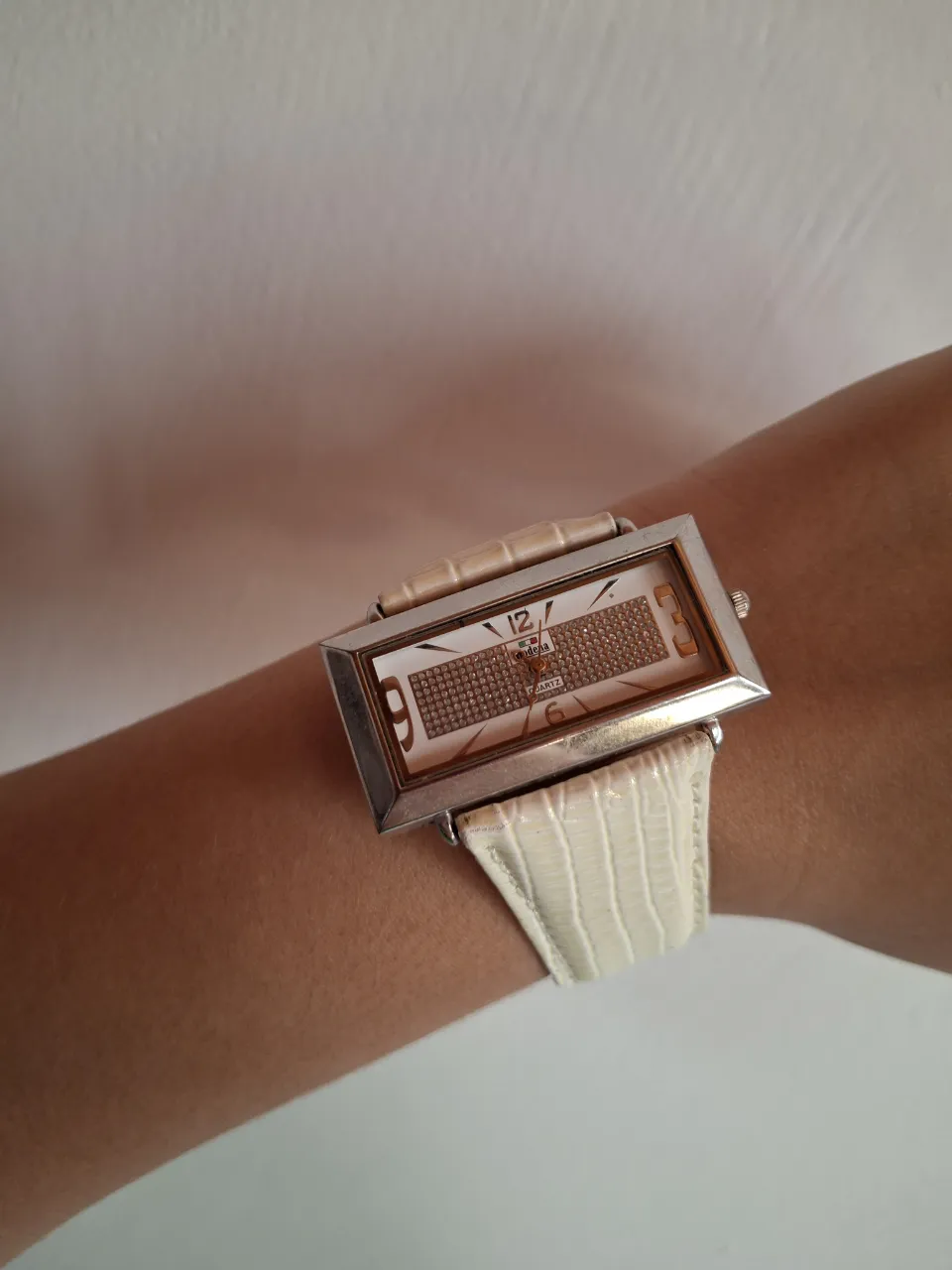 Reloj de pulsera con malla blanca texturizada y caja rectangular plateada. La esfera es blanca con números y detalles en plateado y pequeños cristales incrustados.