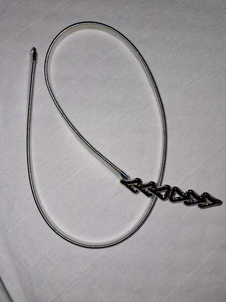 Cinturón fino de metal plateado con cierre de resorte y un dije con forma de flechas o picos.