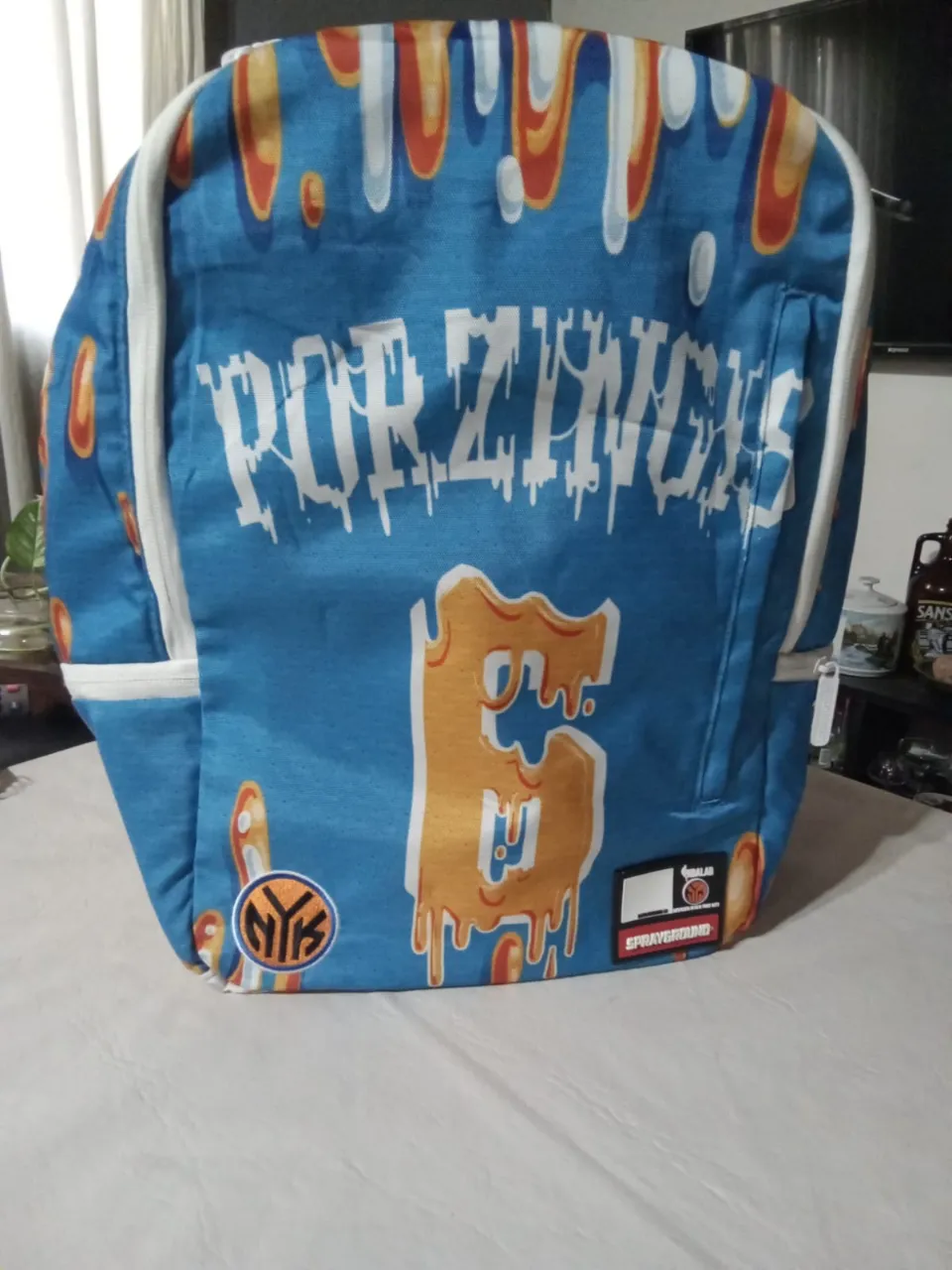NO TIENE DETALLES. 
Mochila azul con estampa de llamas naranjas y blancas. Tiene un número '6' grande en color naranja con efecto de goteo. Incluye parches con logos.

