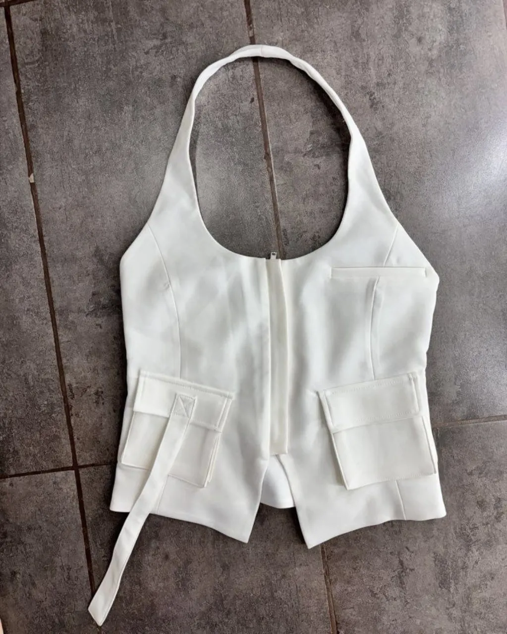 Top blanco con cuello halter, cierre delantero y dos bolsillos con solapa en la parte inferior. Posee un lazo lateral.