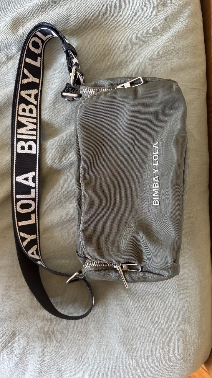 cartera de nylon color verde militar, con correa ajustable estampada con el logo de la marca. Tiene varios compartimentos con cierre.