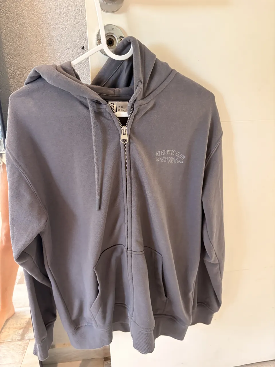 Buzo gris con cierre completo y capucha. Tiene un bordado pequeño en el frente. Ideal para un look casual y deportivo.