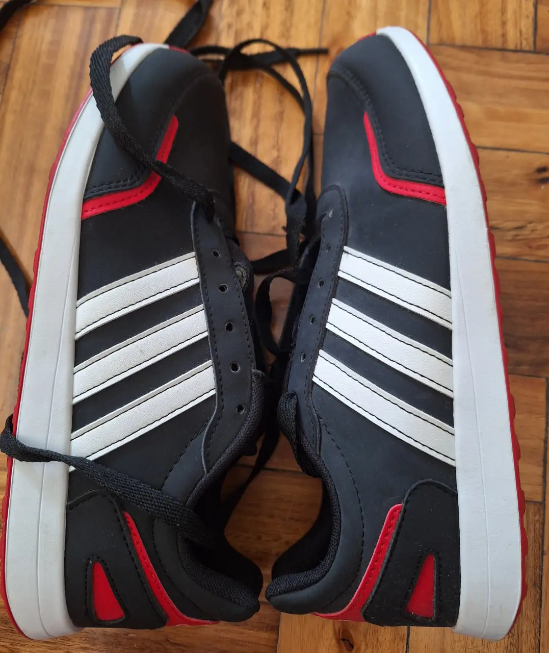 Zapatillas Adidas negras con detalles en rojo y blanco. Diseño deportivo con las icónicas tres rayas blancas. Ideales para uso diario y actividades físicas.