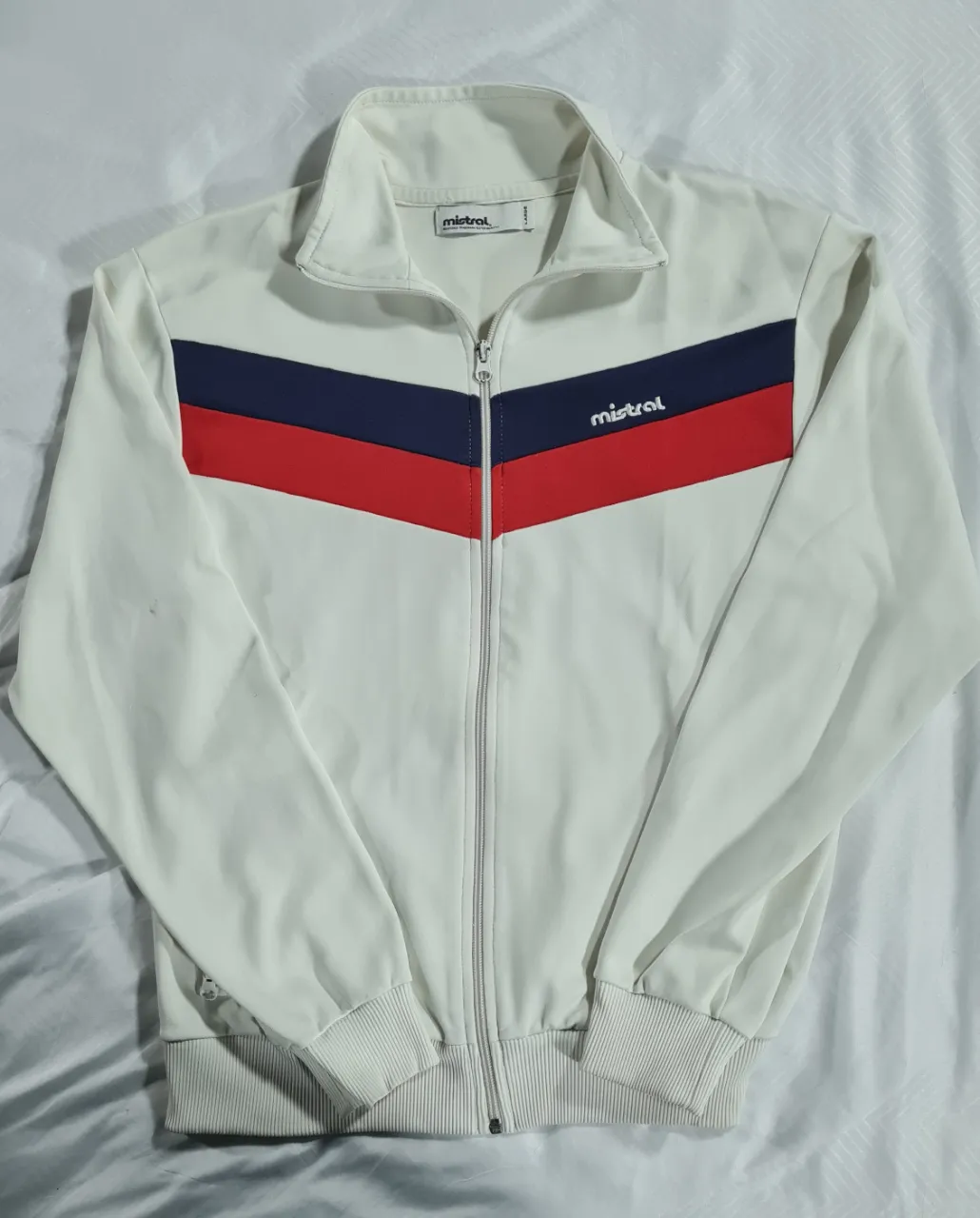 Campera deportiva Mistral blanca con detalles en azul y rojo. Con cierre frontal y cuello alto. Ideal para un look retro y cómodo.
