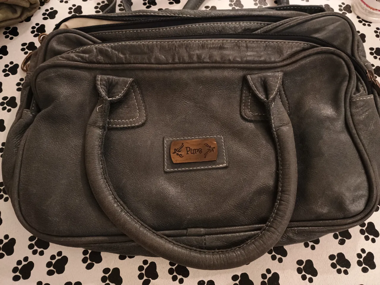 Bolso de mano de cuero gris, con manijas y detalle de etiqueta con logo. Ideal para llevar tus pertenencias con estilo y comodidad.