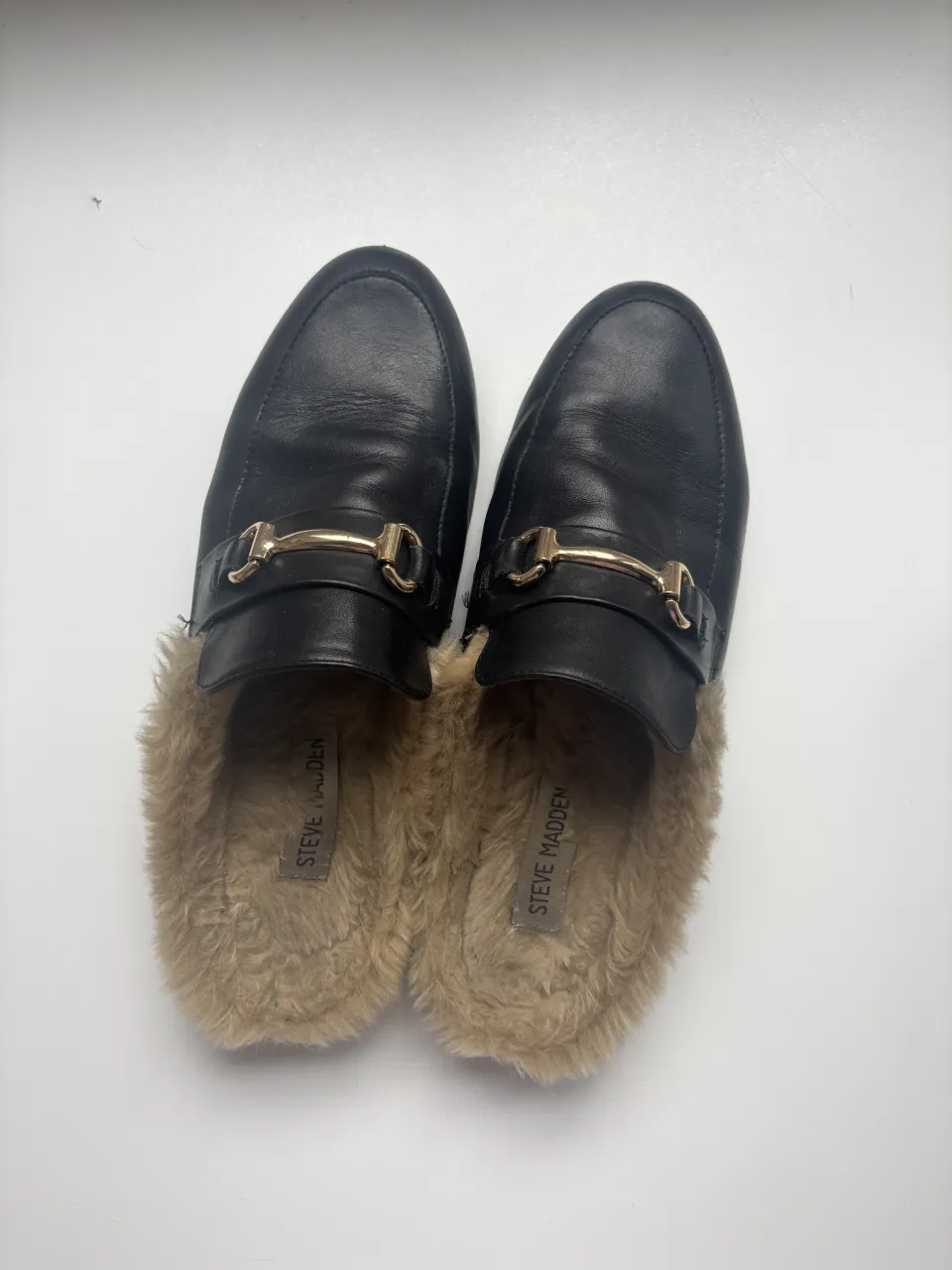 Mocasines negros de cuero con forro de pelo sintético color beige y detalle metálico dorado en la capellada.