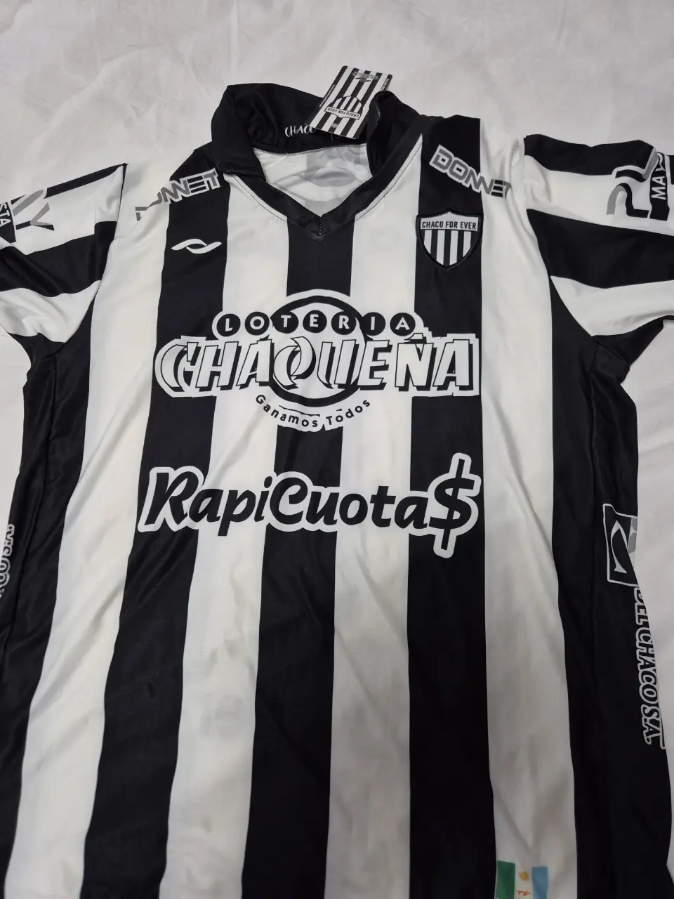 Camiseta de fútbol a rayas blancas y negras del Club Chaco For Ever. Original. Con logos de auspiciantes como Lotería Chaqueña y RapiCuota. Ideal para fanáticos y coleccionistas.