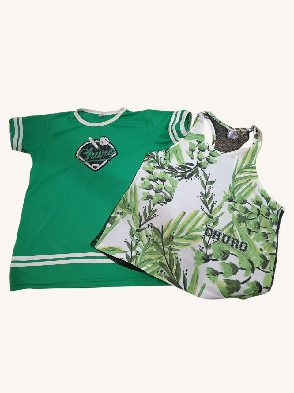 Conjunto de dos remeras deportivas. Una remera verde con mangas cortas y detalles a rayas en los puños y el bajo, con un logo de béisbol bordado en el frente. La otra es una musculosa blanca con estampa de hojas verdes y la palabra "CHURO" estampada. La parte trasera de la musculosa es de red negra.
‼️Detalle en la remera: una mancha.