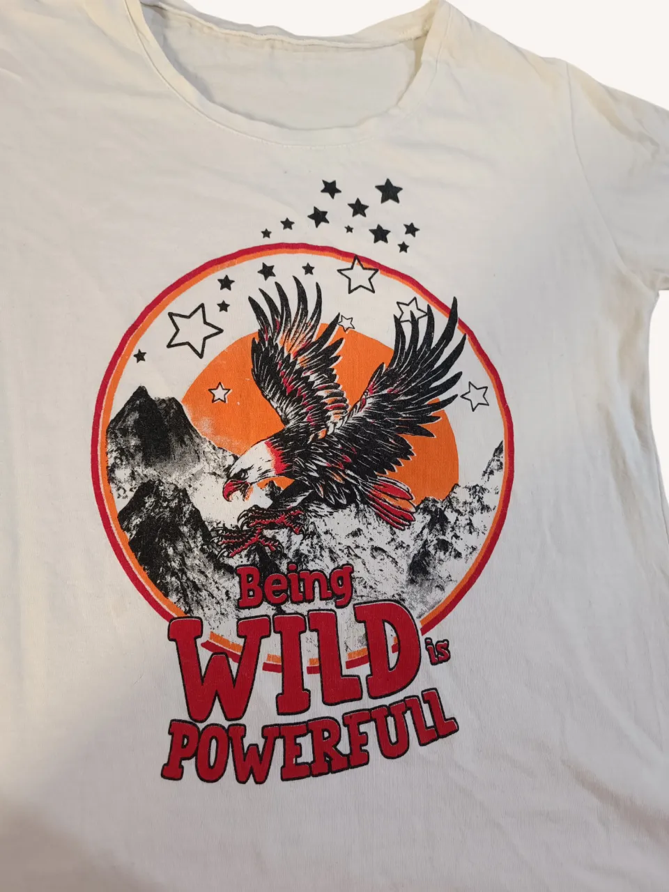 Remera blanca con estampa de un águila en vuelo sobre montañas y el texto "Being WILD is POWERFULL". La estampa tiene un círculo naranja de fondo y estrellas negras y blancas.
📏Ancho 41 cm.
