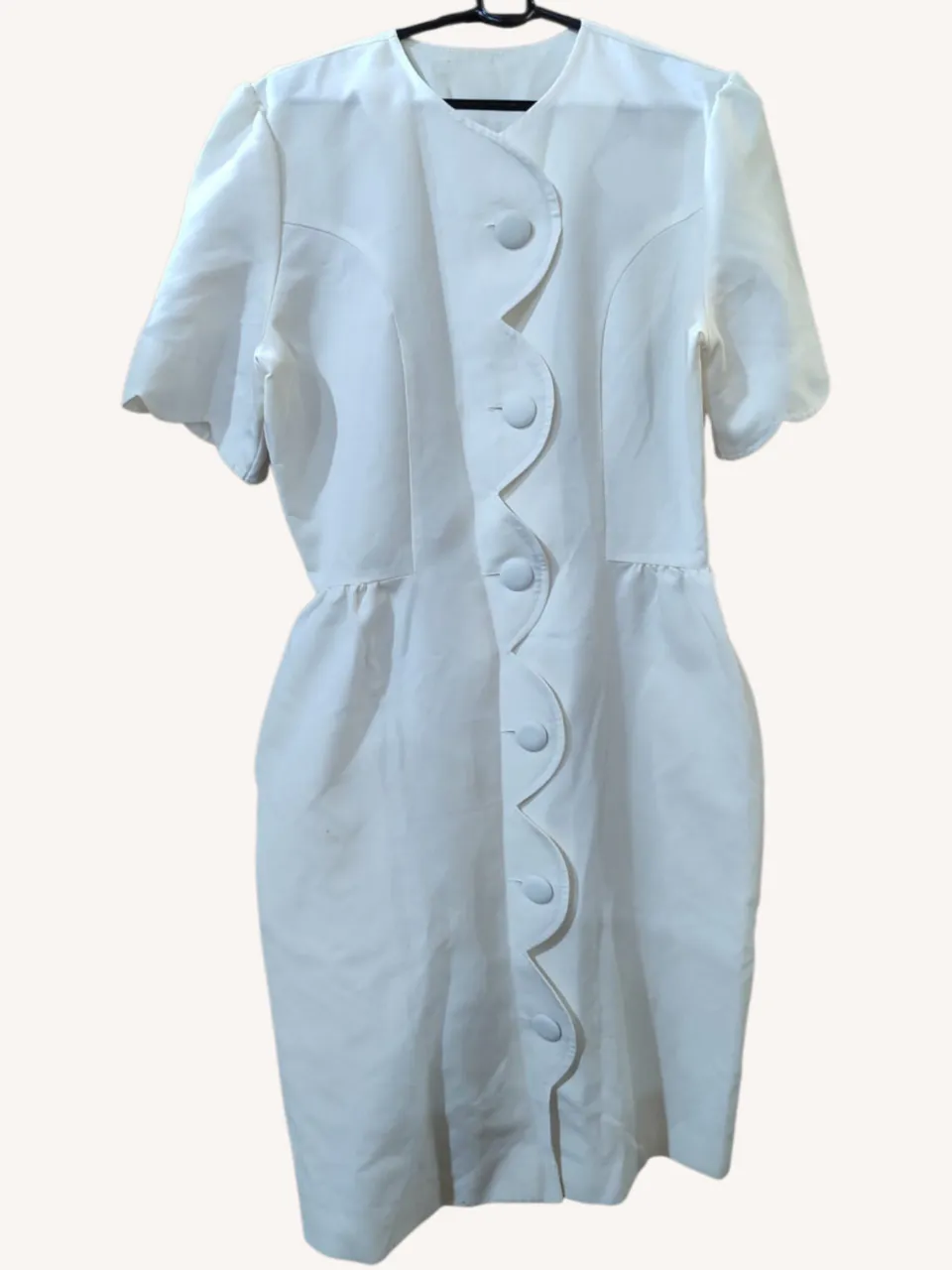 Vestido vintage blanco de corte recto, con mangas cortas y escote redondo. Posee un diseño de botones curvos en el frente y detalles de frunces en la cintura. Ideal para un look elegante y sofisticado. Tiene bolsillos amplios.