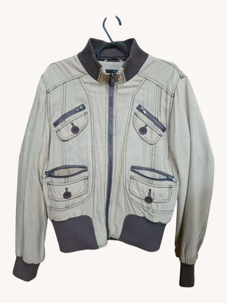 Campera bomber color beige efecto desgastado con detalles en marrón. Posee cierre central, cuello y puños de ribb. Tiene bolsillos con solapa y botones, y bolsillos con cierre.