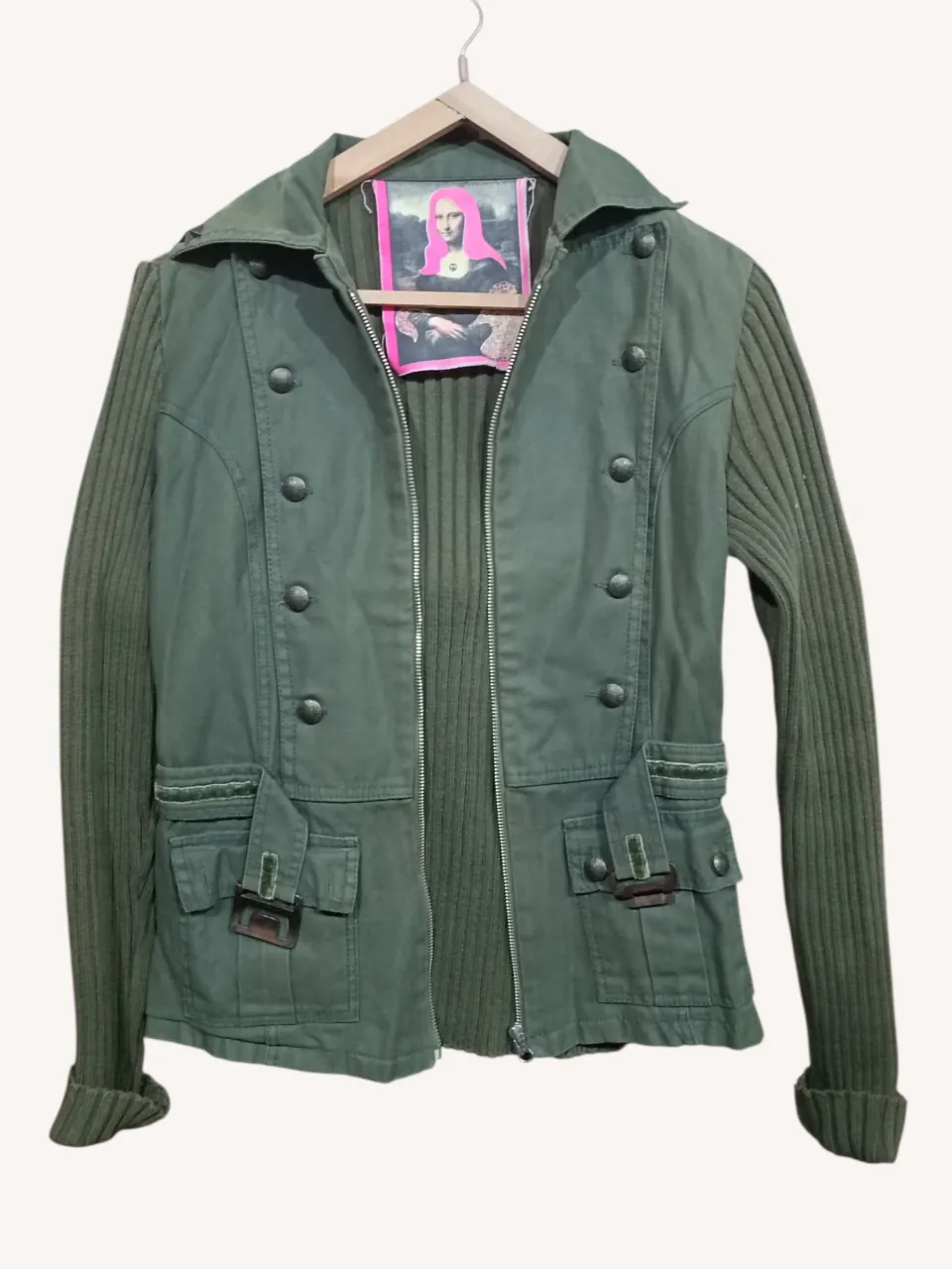 Campera verde militar con mangas de tejido acanalado. Posee doble hilera de botones al frente, cuello con solapa y detalle de estrella metálica. Incluye cinturón de tela con hebilla metálica y bolsillos con solapa en la parte inferior. El interior tiene un estampado de camuflaje.
‼️Detalle: decoloración en el cuello.
