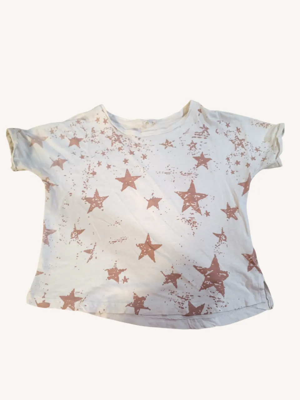 Remera blanca con estampa de estrellas en color rosa pálido y salpicaduras. Tiene mangas cortas y cuello redondo.
‼️ Desgaste en una manga.