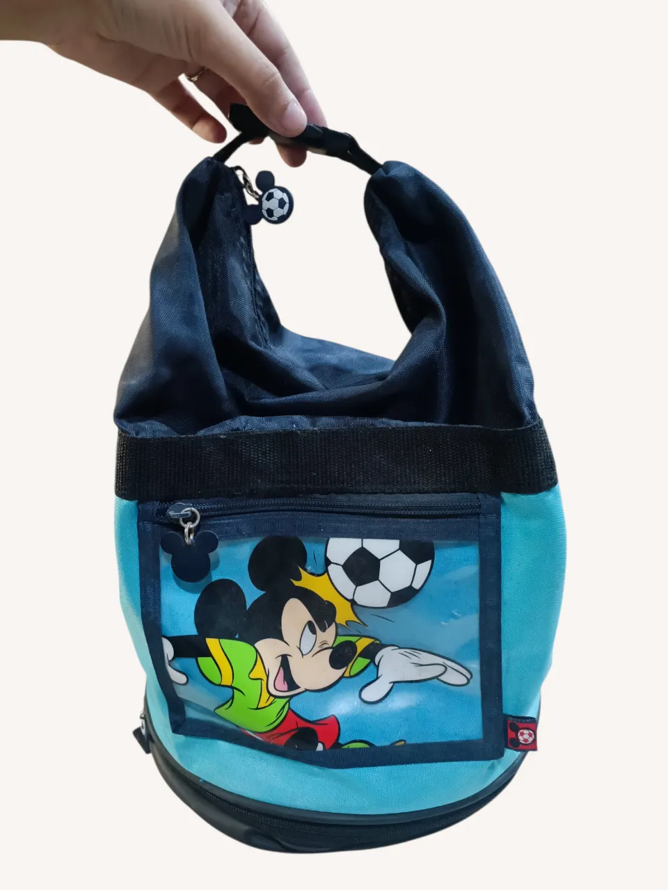 Mochila infantil azul y celeste con estampa de Mickey Mouse jugando al fútbol. Tiene un bolsillo frontal transparente con cierre, compartimento lonchera en la base y tirador de Mickey, y tiras para colgar.