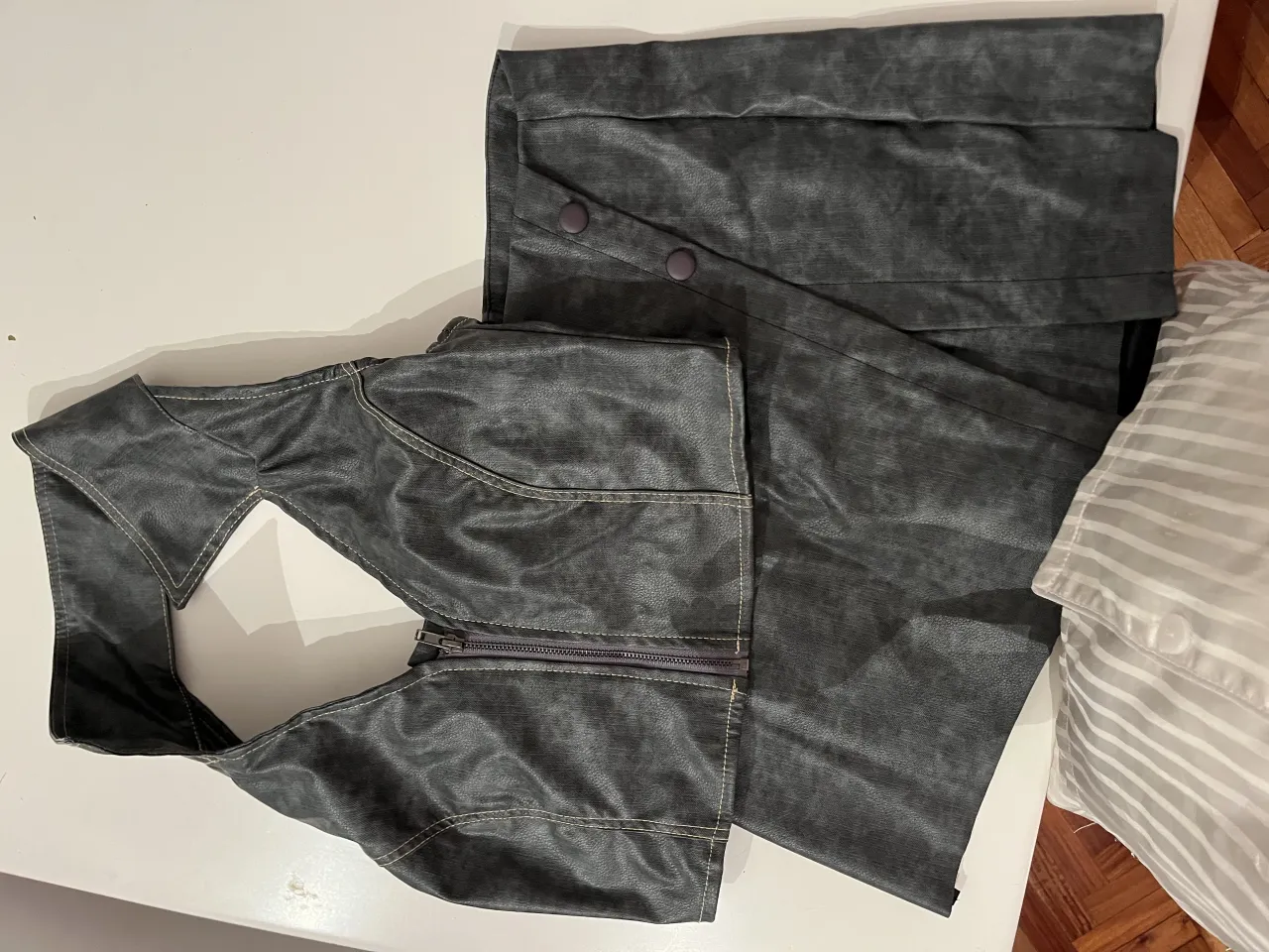 Conjunto de falda y top estilo, ambos de cuero sintético gris. La falda es corta, con un diseño plisado y un cierre en la parte trasera. El top es tipo chaleco, con cuello solapa, cierre delantero y detalles de costura visibles. Ideal para un look audaz y moderno.