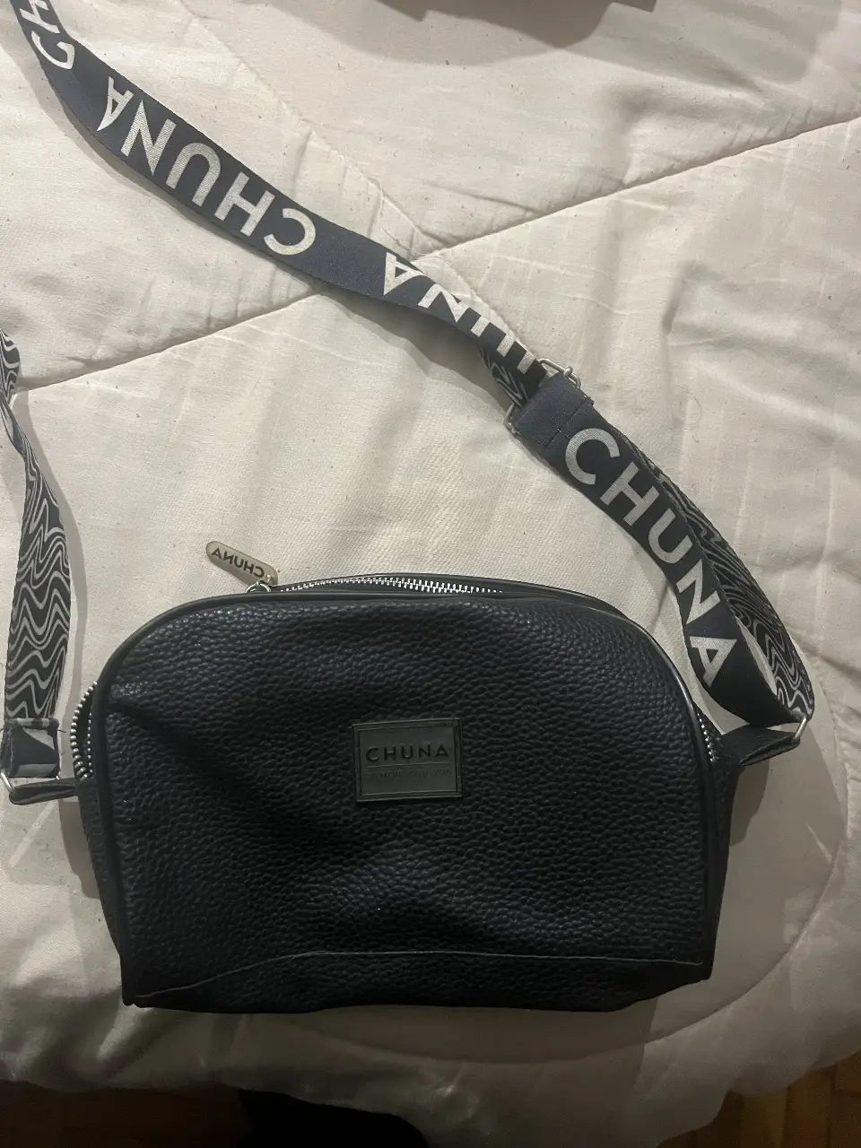 Bandolera negra de cuero sintético con correa ajustable y detalle de la marca CHUNA. Ideal para llevar lo esencial con estilo y comodidad.