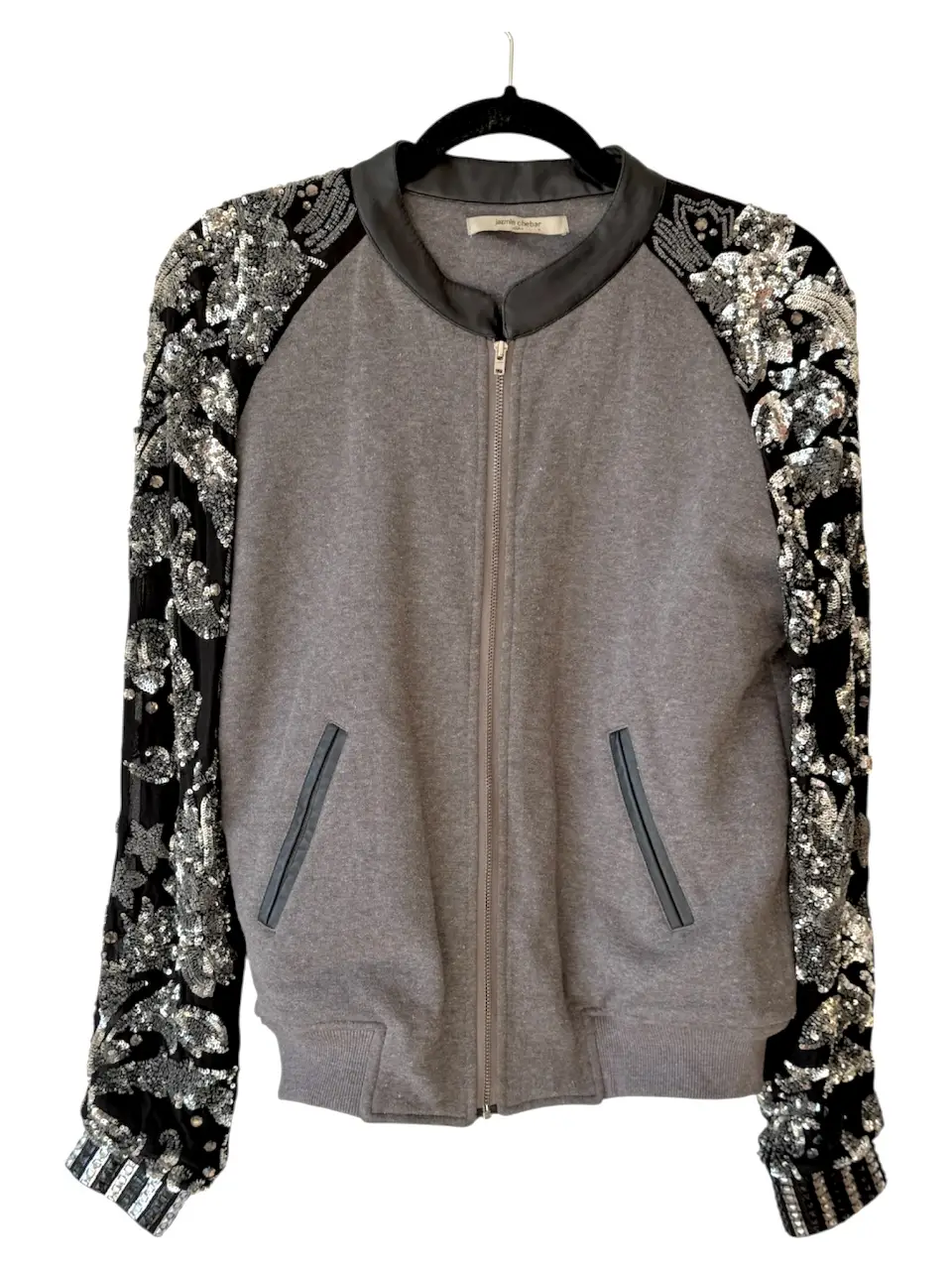 Campera bomber gris con mangas negras bordadas con lentejuelas plateadas. Cuello y bolsillos con detalles en cuero negro. Cierre frontal.