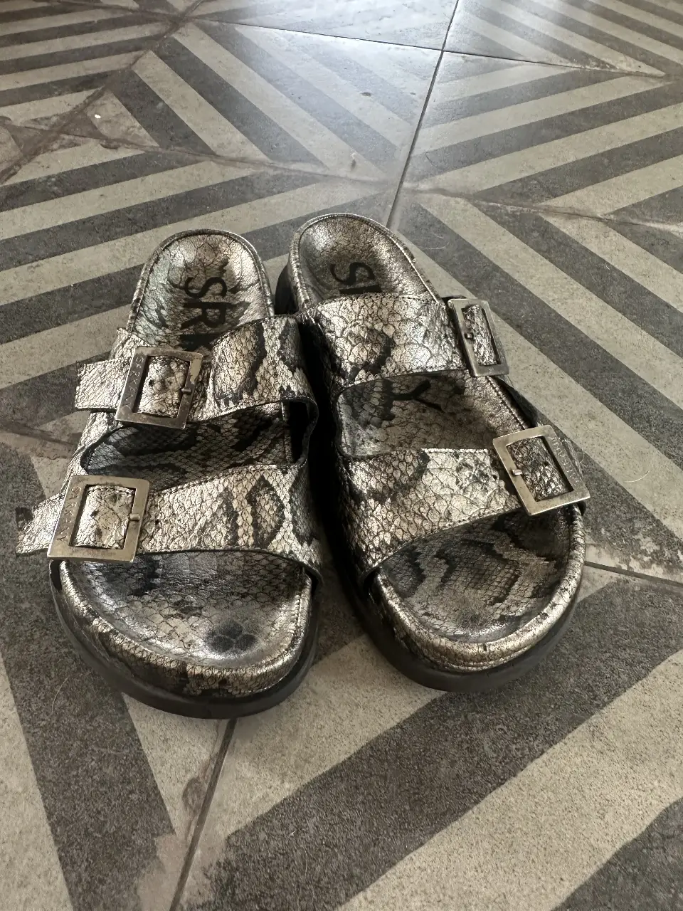 Sandalias de mujer con diseño de serpiente en tonos grises y plateados. Cuentan con dos hebillas ajustables y suela negra. Ideales para un look casual y moderno.