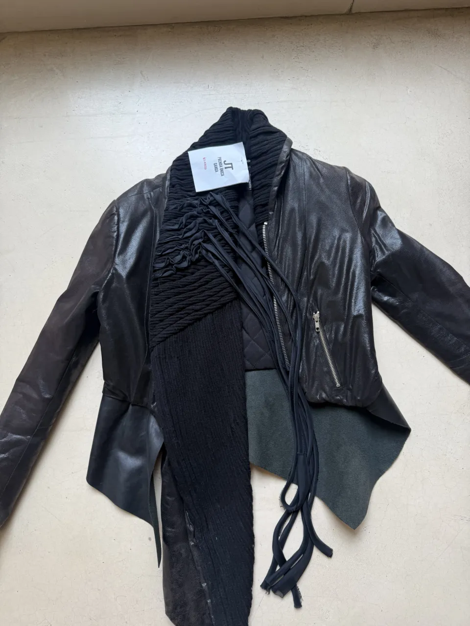 Campera de cuero negra con un diseño vanguardista y desestructurado. Presenta detalles de fruncidos y tiras colgantes que le dan un toque único y moderno. Ideal para quienes buscan un look audaz y con personalidad.