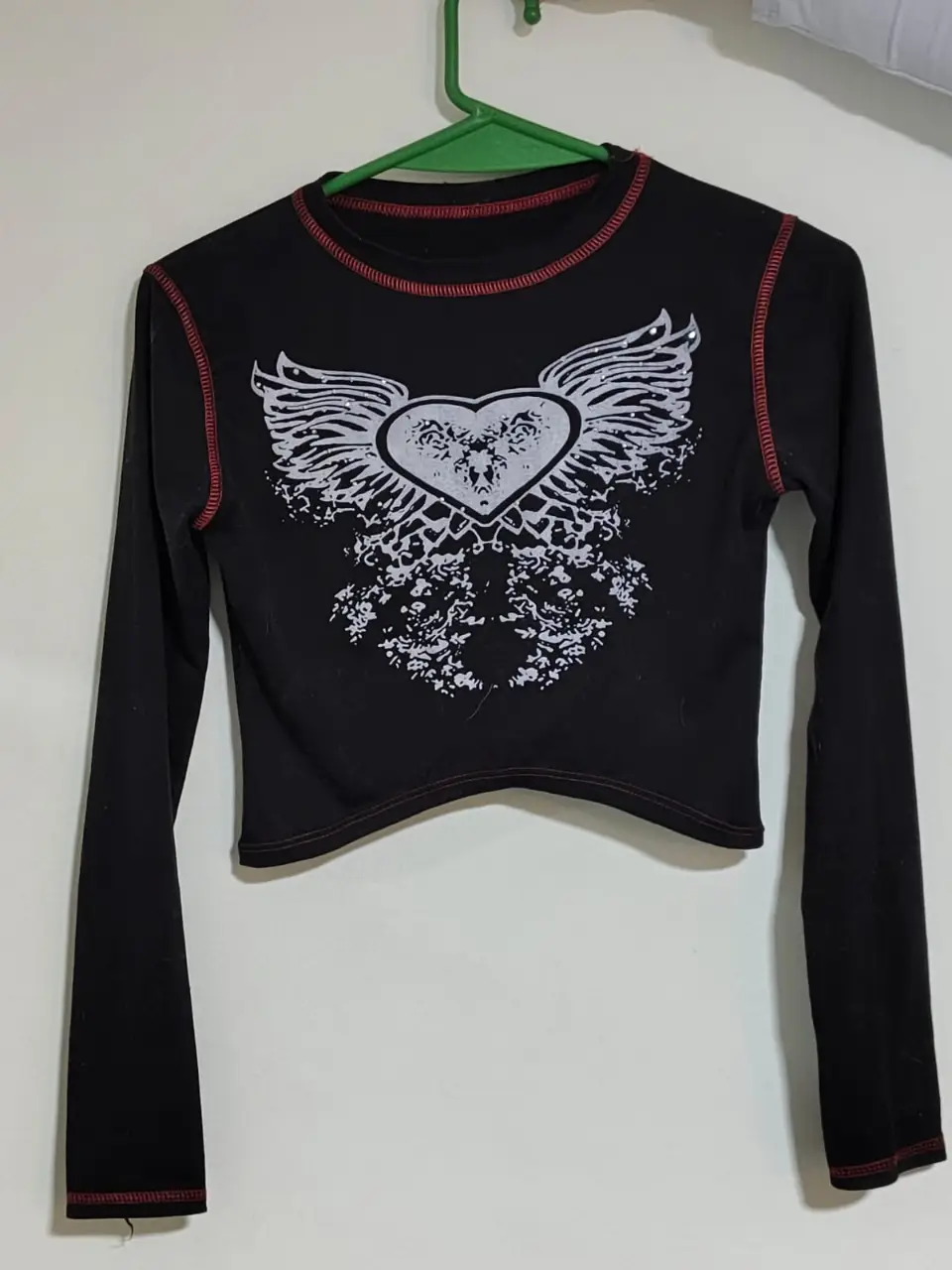 Remera manga larga negra con estampado de corazón alado en blanco y costuras rojas a la vista. Ideal para un look Y2K o rocker. MEDIDAS: LARGO 34//ANCHO 33//MANGA: 57//