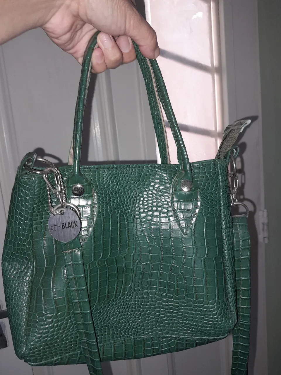 Bolso de cuero verde con textura de cocodrilo. Tiene dos asas cortas y una correa larga desmontable. Incluye un dije metálico con la inscripción "off - BLACK".