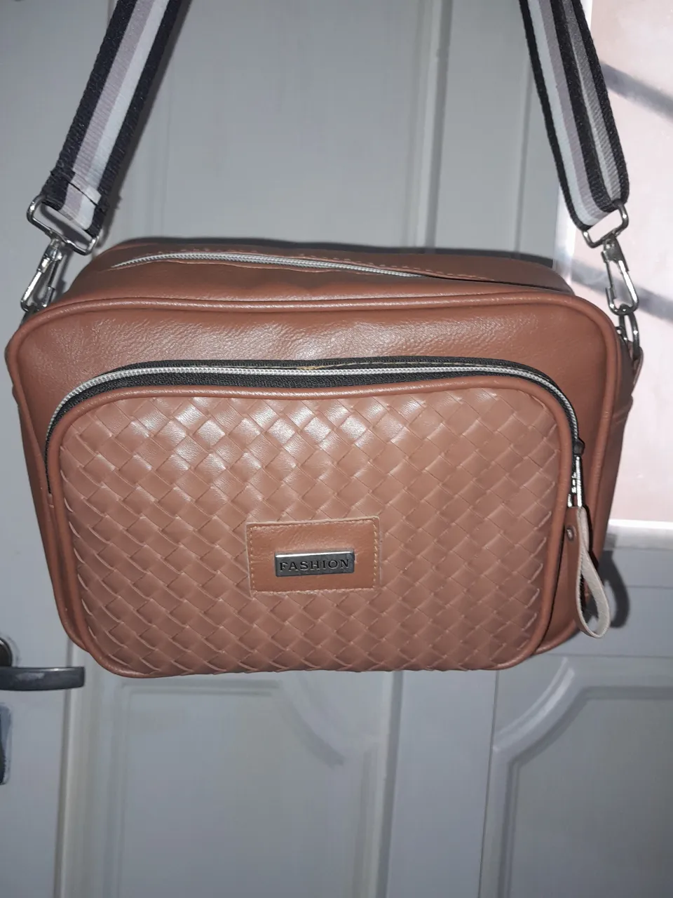 Bolso marrón con correa de tela a rayas blancas, negras y grises. Tiene un bolsillo frontal tejido con un detalle metálico con la palabra "FASHION".