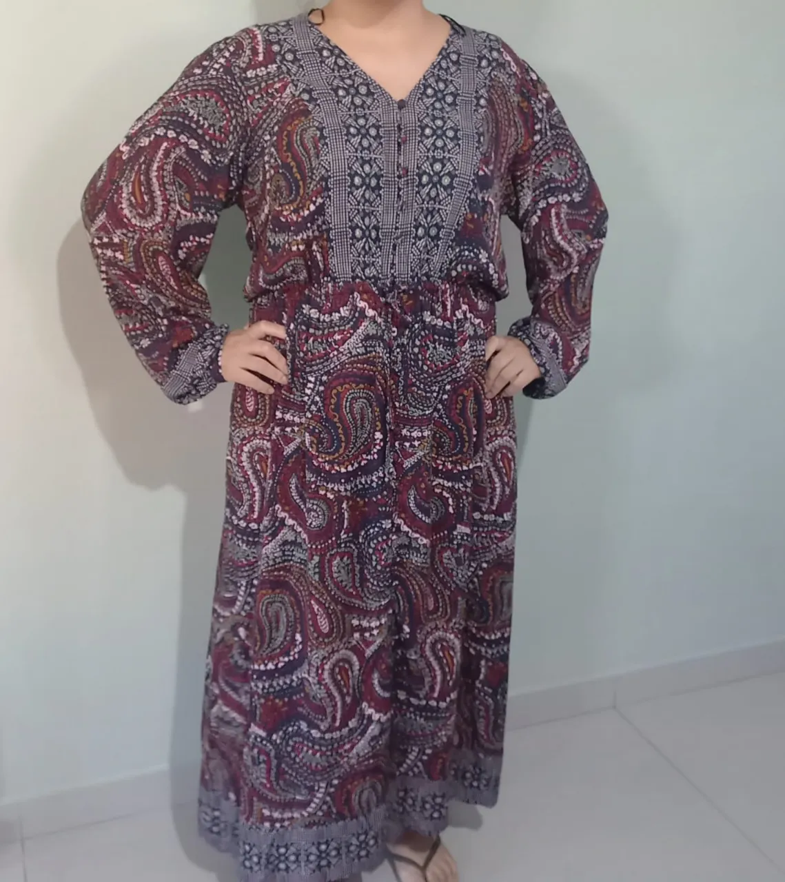 Vestido largo estampado con mangas largas y escote en V. Posee detalles de bordado en la parte superior y un cordón para ajustar en la cintura. El estampado es de estilo paisley en tonos bordó, negro, blanco y verde.