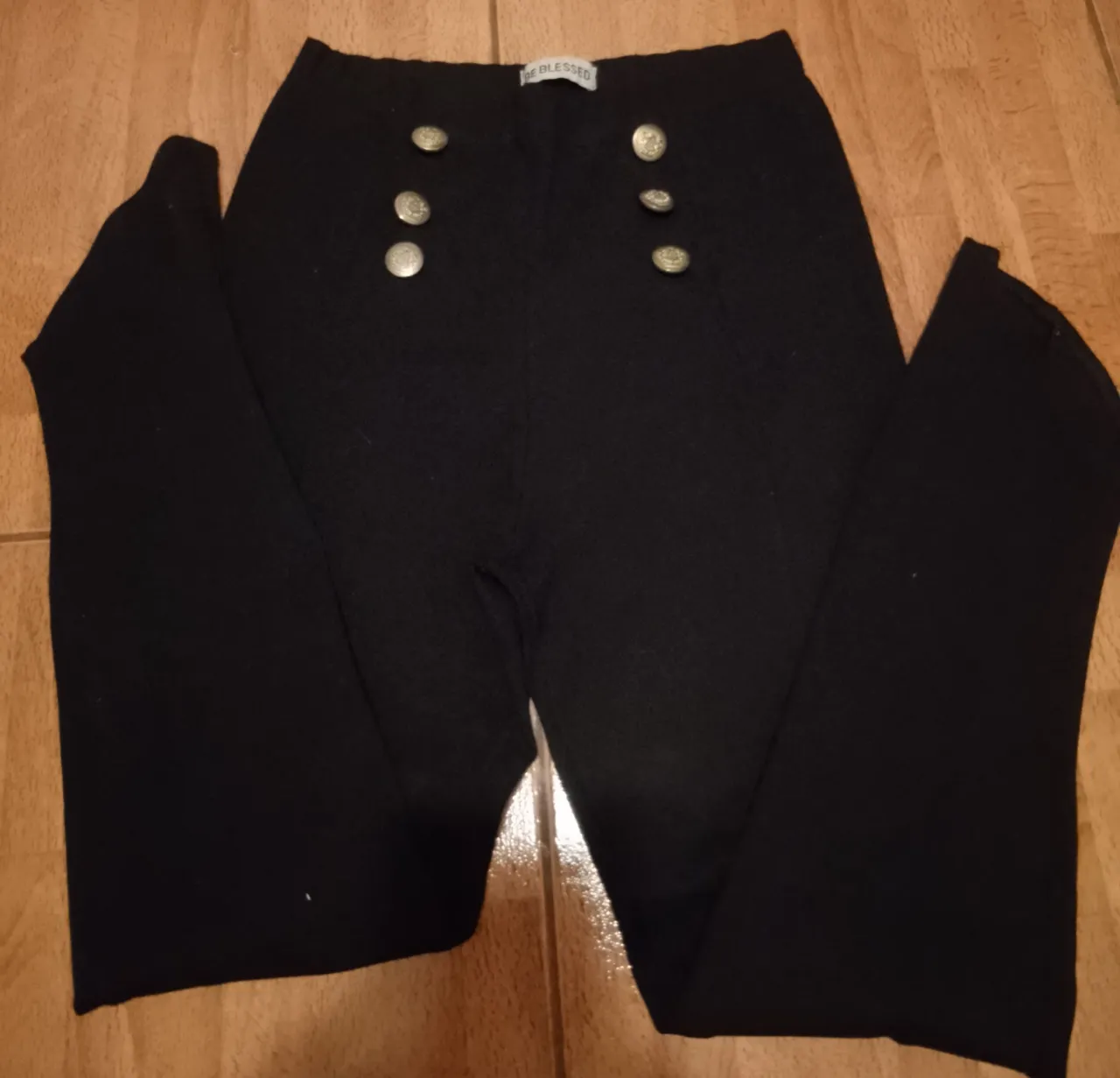 Pantalón negro con seis botones dorados en la parte delantera. Etiqueta interior con la marca 'BE BLESSED'.