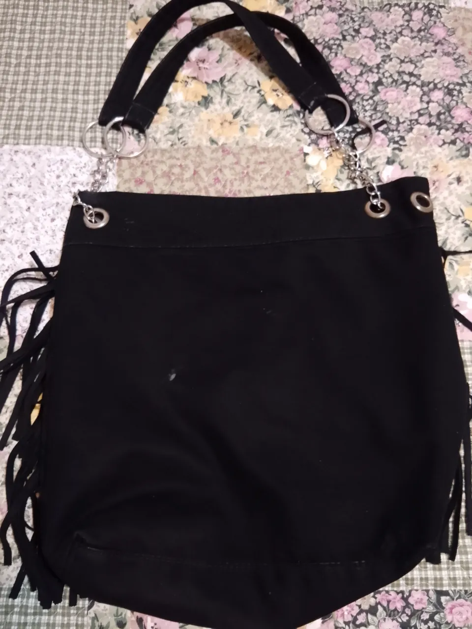 Cartera negra gamuzada con flecos largos en los laterales y en la parte inferior. Posee dos asas negras y detalles de cadena y argollas metálicas. El interior cuenta con un bolsillo con cierre.