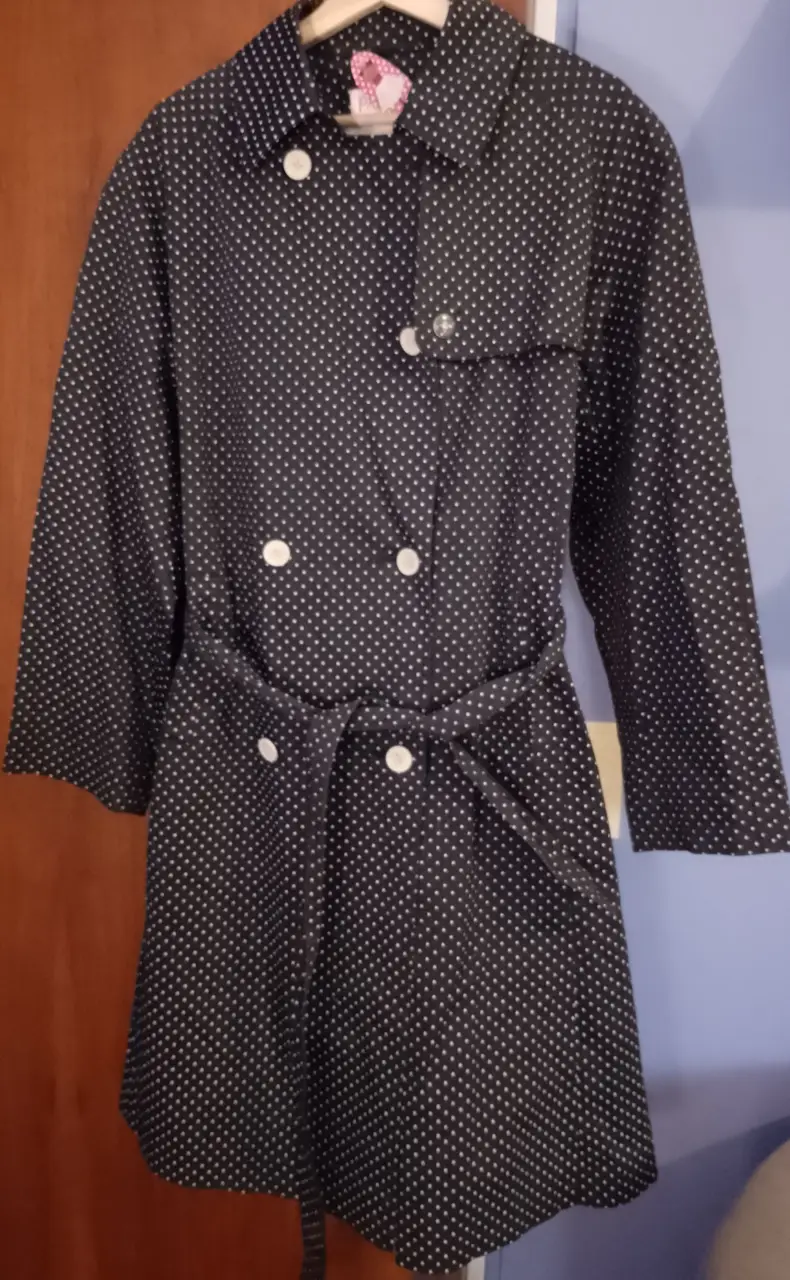 Sobretodo de mujer con estampado de lunares blancos sobre fondo negro. Diseño con botones, cinturón para ajustar la cintura y forro interior blanco. Ideal para un look vintage y elegante.