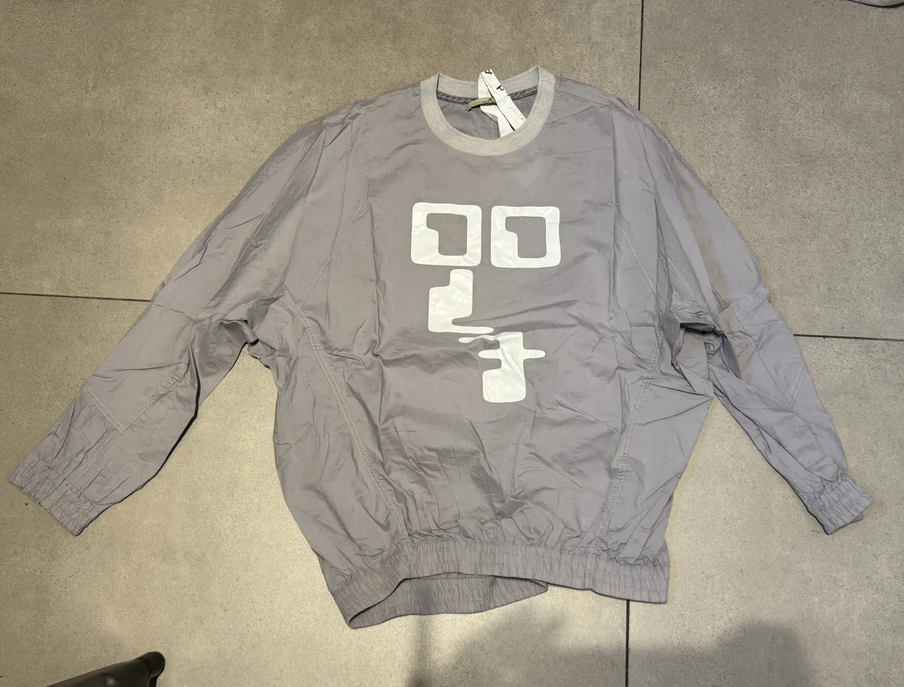 Remera gris con estampa blanca en el frente. La estampa tiene un diseño abstracto con formas cuadradas y líneas. En la espalda, tiene una etiqueta con texto y un diseño de cuadros pequeños.