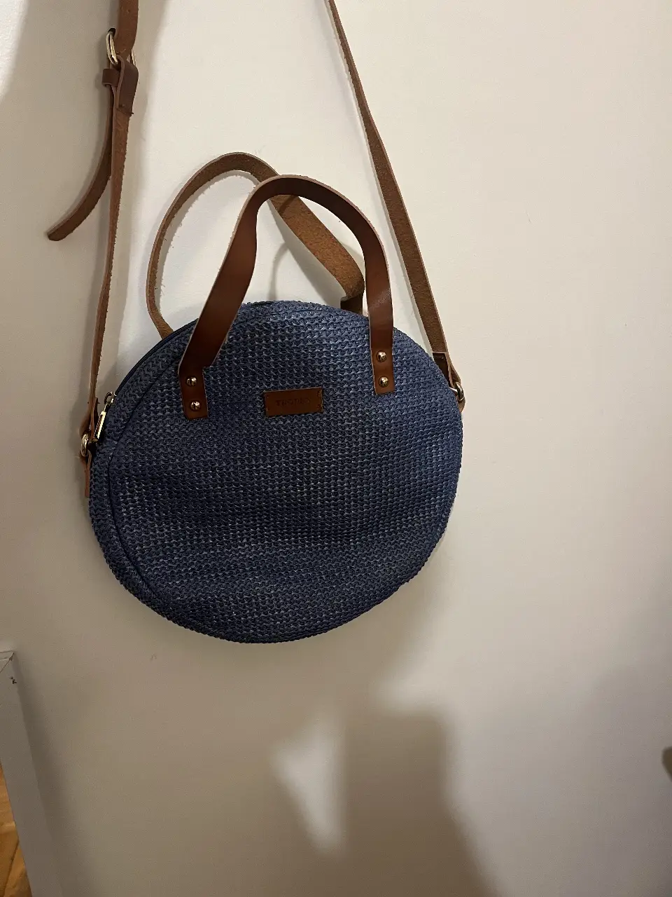 Cartera redonda de mujer, azul con detalles en marrón. Confeccionada en material tejido, con manijas y correa para colgar al hombro. Interior estampado con la marca Tropea.