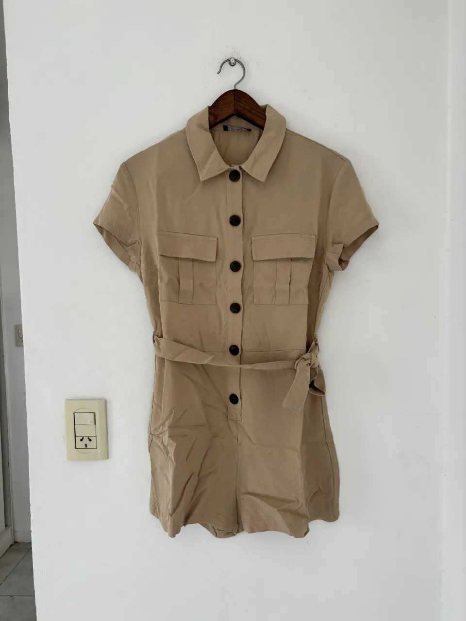 Mono corto de color beige con cuello camisero, mangas cortas y cinturón de tela. Posee dos bolsillos superiores con solapa y cierre de botones al frente.