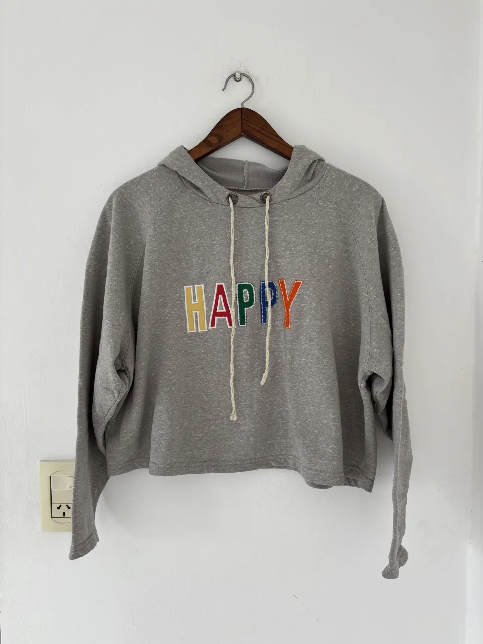 Buzo gris con capucha y cordones blancos. En el frente, la palabra "HAPPY" está bordada con letras de colores: amarillo, rojo, verde, azul y naranja.