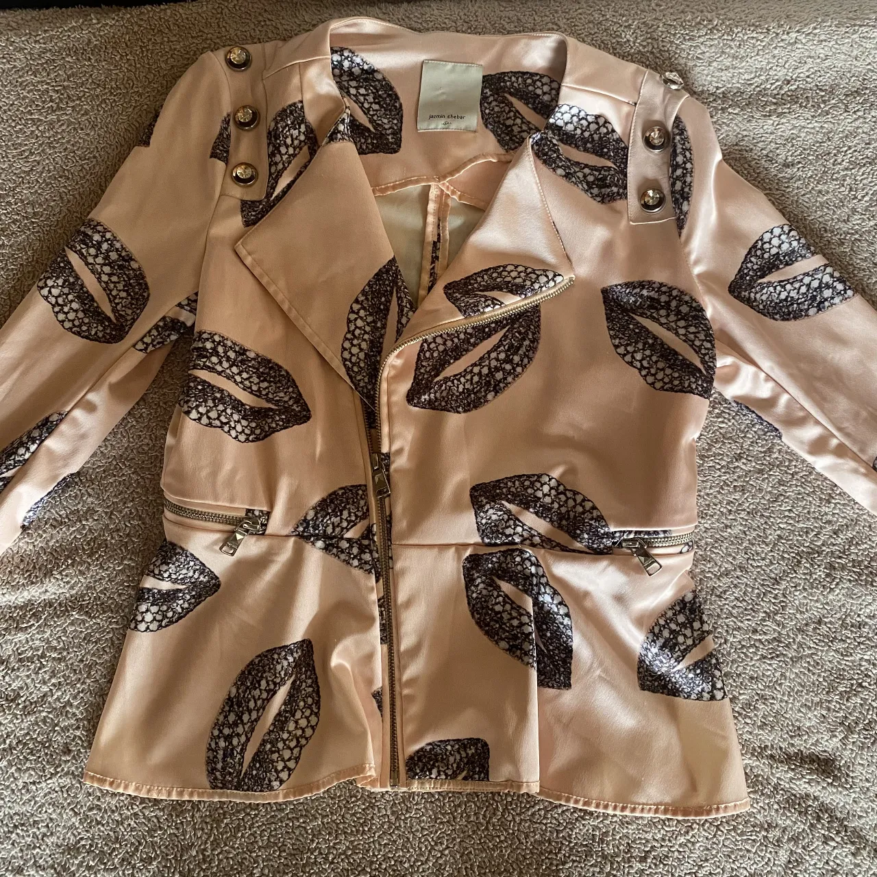 Blazer rosa pálido con estampa de labios en negro y blanco. Diseño tipo motoquero con cierre metálico al frente y en los bolsillos. Detalle de botones dorados en los hombros. Una prenda única y audaz para destacar tu estilo.