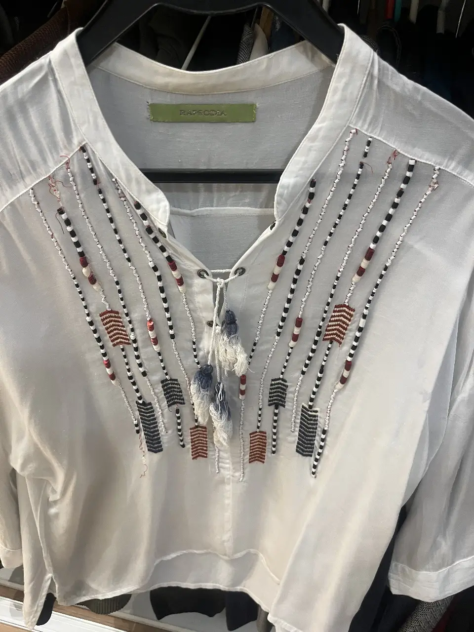 Blusa blanca con detalles bordados en azul y bordo y borlas en el cuello. Estilo boho chic, ideal para un look casual y fresco.