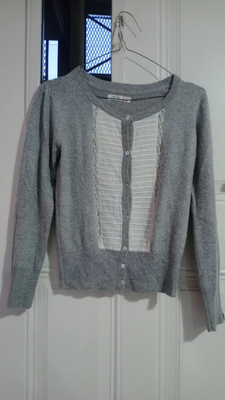 Cardigan gris con botones al frente y detalles de encaje y tela blanca plisada en el frente. Mangas largas y cuello redondo.