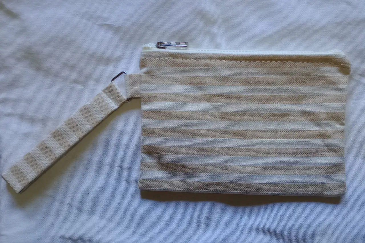 Monedero con diseño a rayas en tonos crudo y marrón. Cuenta con un cierre y una manija para llevarlo cómodamente. Ideal para guardar monedas, tarjetas o pequeños objetos.