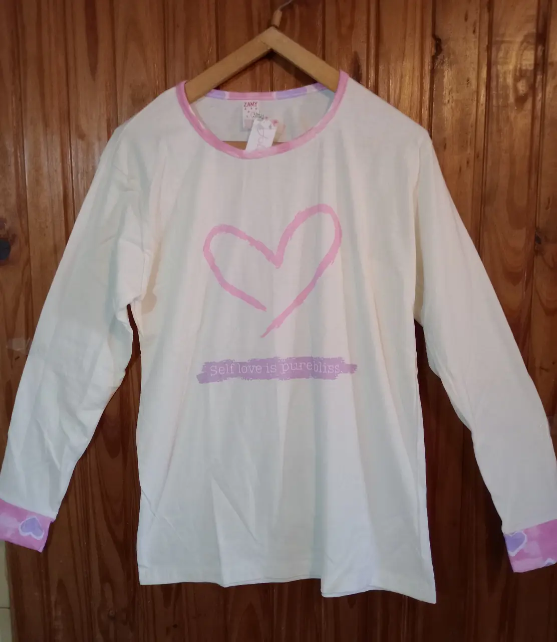 Conjunto de pijama de mujer, con remera manga larga blanca con estampado de corazón rosa y frase "Self love is pure bliss". Pantalón largo rosa con estampado de corazones.