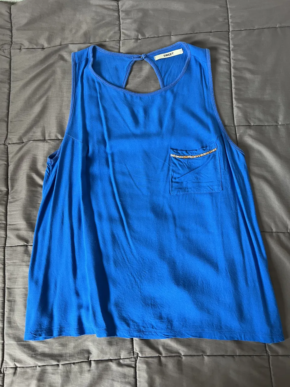 Musculosa azul con bolsillo en el frente. El borde del bolsillo tiene detalles de pedrería dorada y naranja. En la espalda tiene un recorte en forma de gota con un botón.