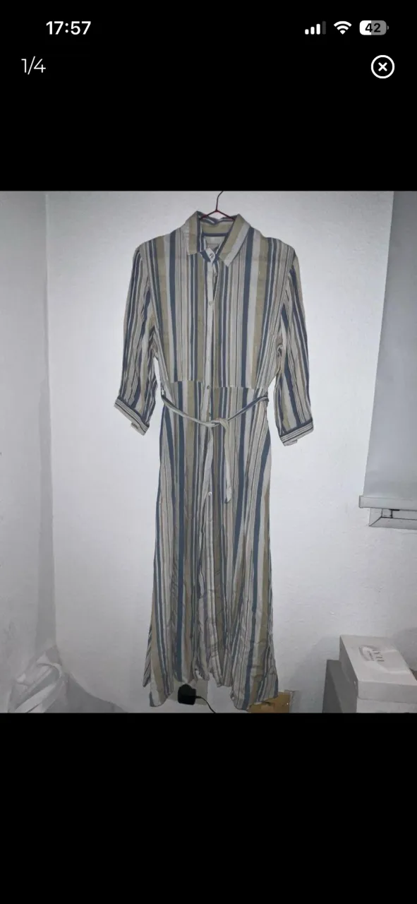 Vestido largo a rayas verticales en tonos blanco, azul y crudo. Posee cuello de camisa, manga 3/4 y cinturón de tela a tono.